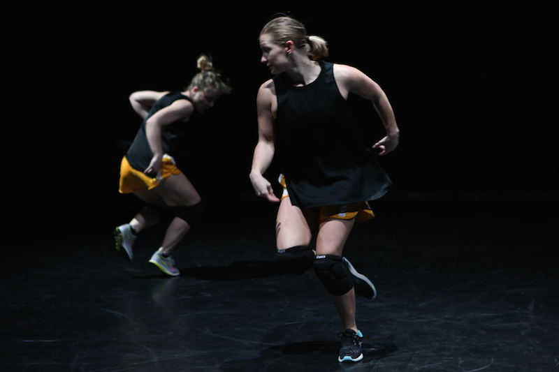 Two in mid run. Their legs are deeply bent. One looks over her shoulder. view is in profile. She wears a black tank top, orange athletic shorts, knee pads and sneakers.